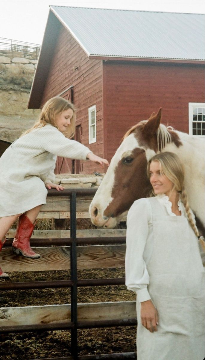 a woman and girl petting a horse on the nose in front of a barn