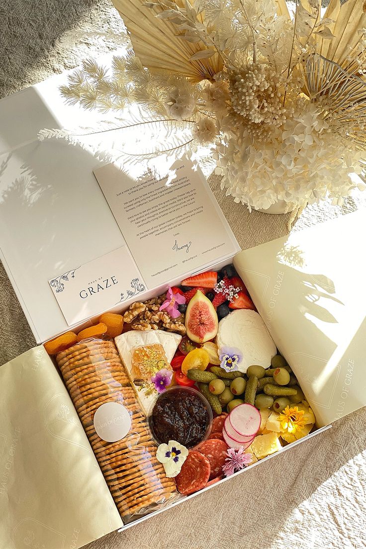 an open box filled with assorted food on top of a white blanket next to a vase