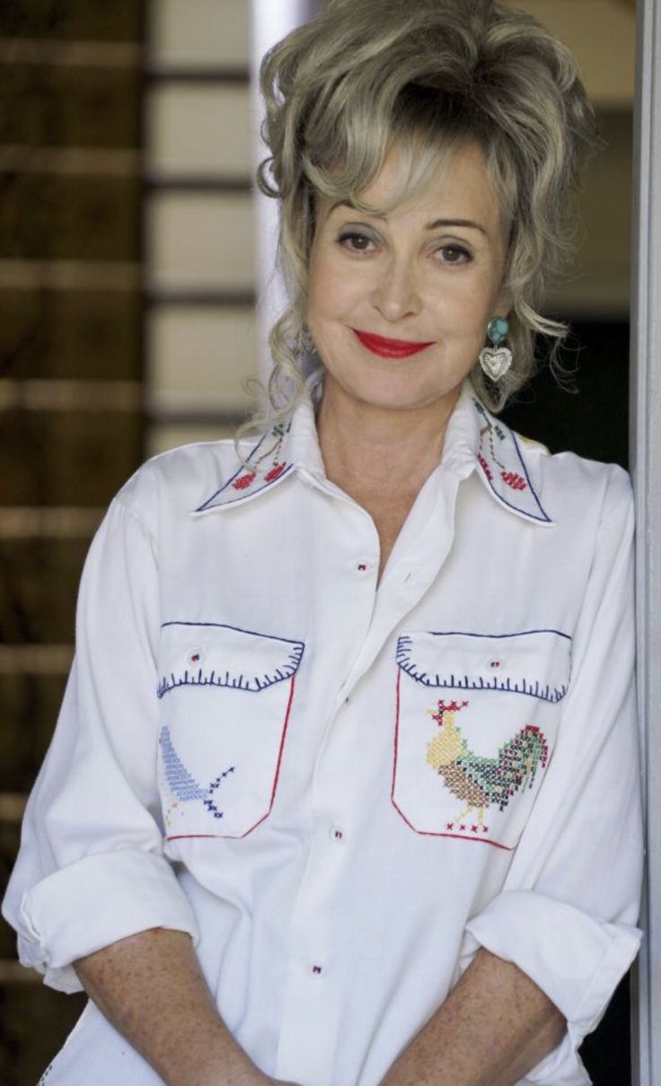 an older woman with grey hair wearing a white shirt and red lipstick, leaning against a wall