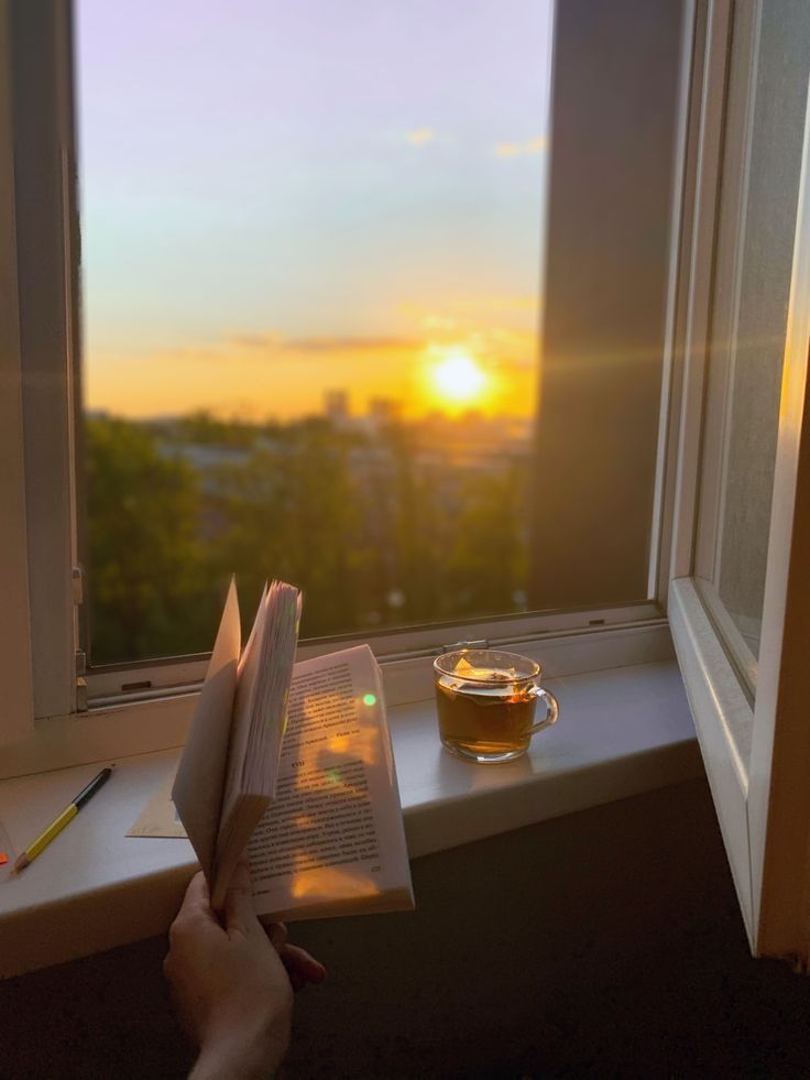 a cup of tea and book on a window sill with the sun setting in the background