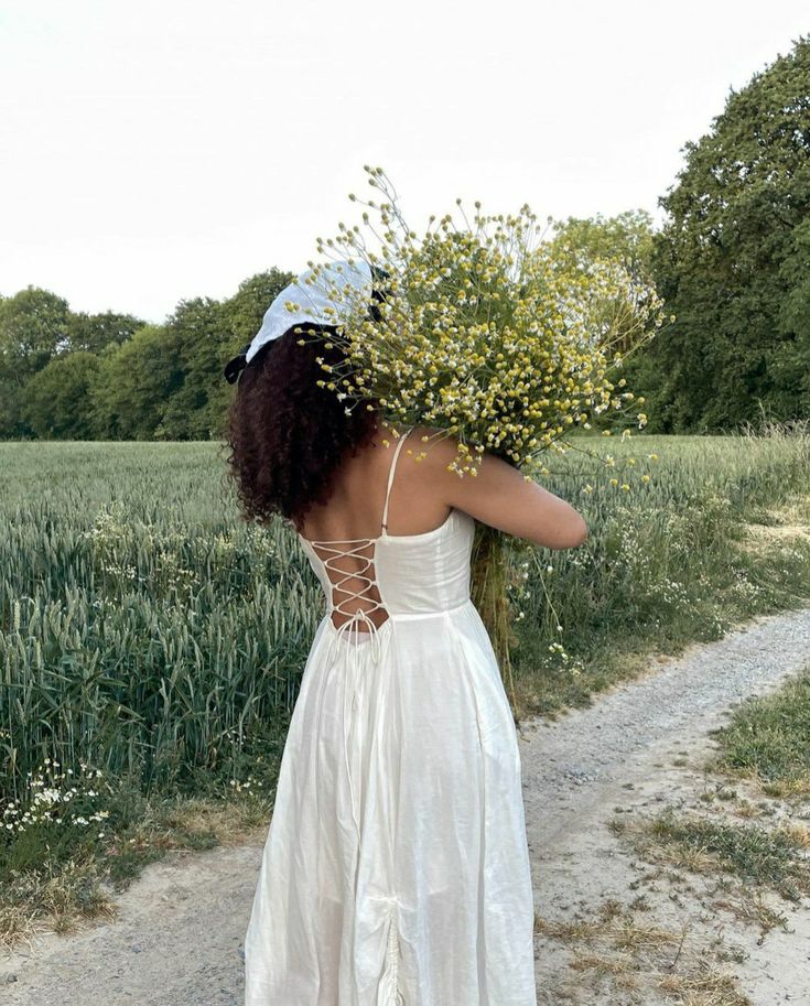 a woman walking down a dirt road holding a bunch of flowers in her hands and wearing a white dress