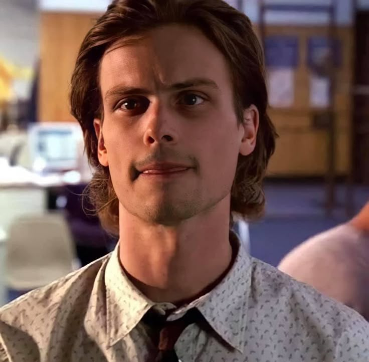 a man with long hair wearing a shirt and tie looking at the camera while standing in an office