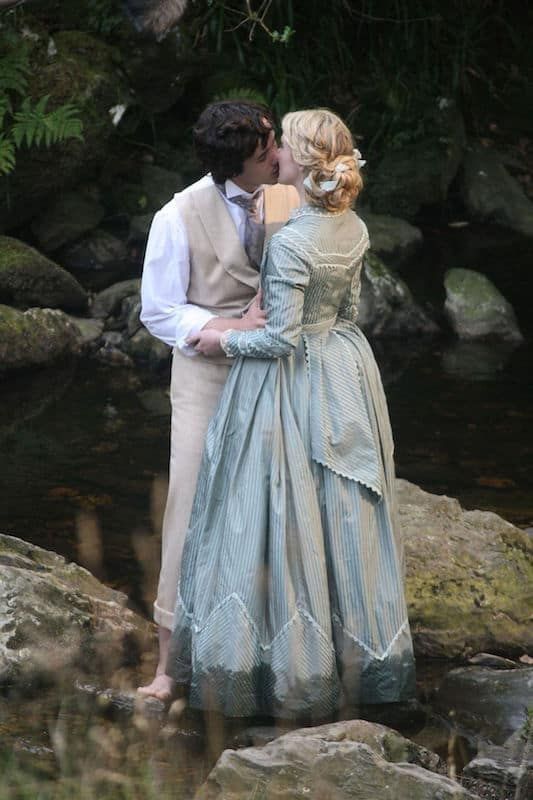 a man and woman dressed in period clothing kissing on rocks next to a river with trees