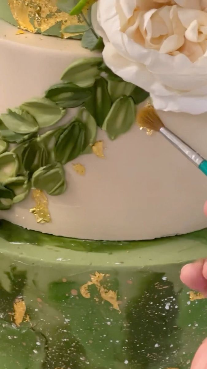 a person painting a white flower on a green cake with gold flakes and leaves