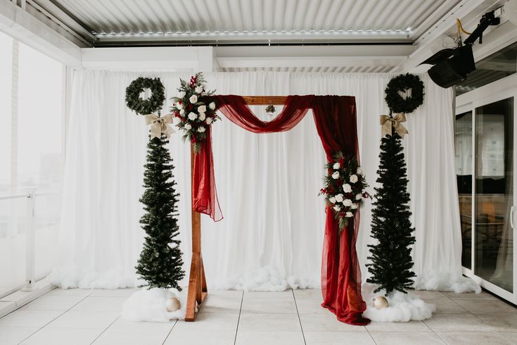 a white and red wedding arch decorated with wreaths, flowers and pine trees for the ceremony