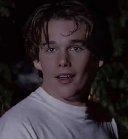 a young man in white shirt standing next to green plants and trees with his eyes wide open