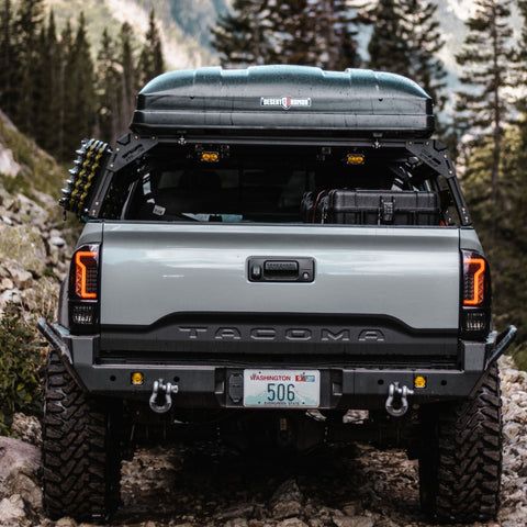 the back end of a silver truck parked on top of a rocky hill with trees in the background