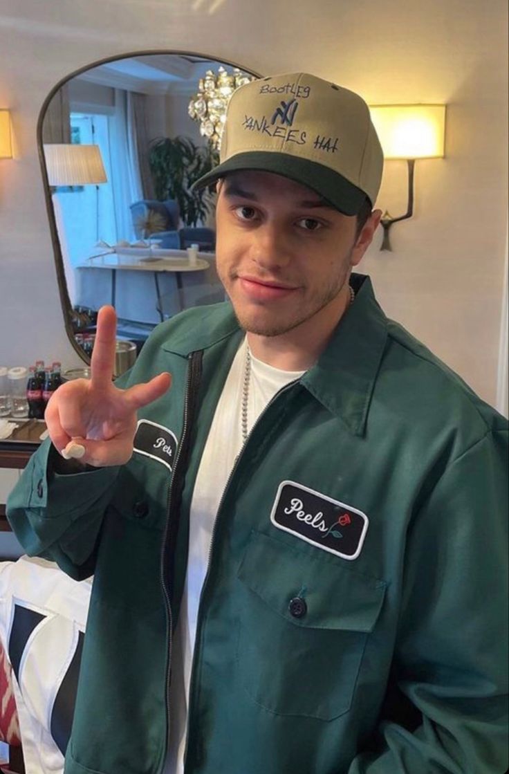 a man wearing a green jacket and hat making the peace sign with his hand while standing in front of a mirror