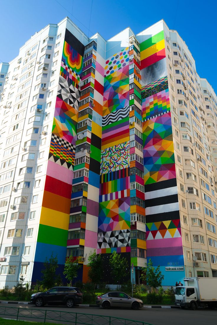 a large multicolored building with cars parked in front of it and tall buildings behind it