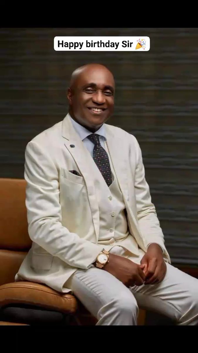 a man in a white suit and tie sitting on a chair with his hands folded