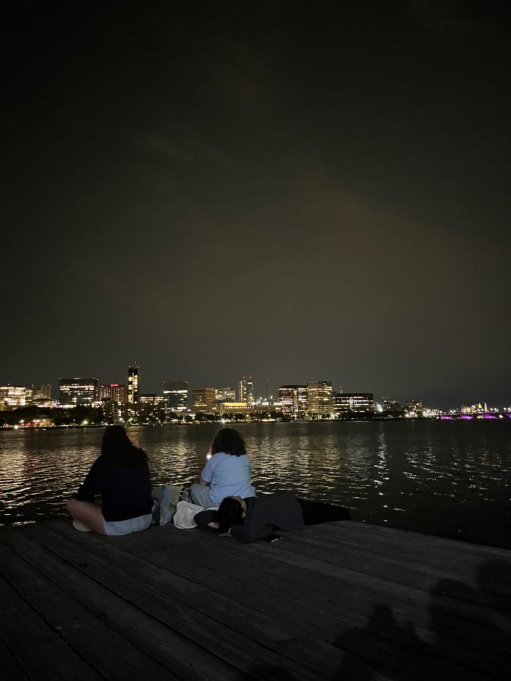 two people sitting on a dock looking at the city lights from across the water in front of them