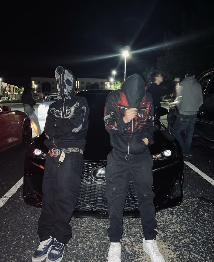 two young men sitting on the hood of a black sports car in front of some parked cars