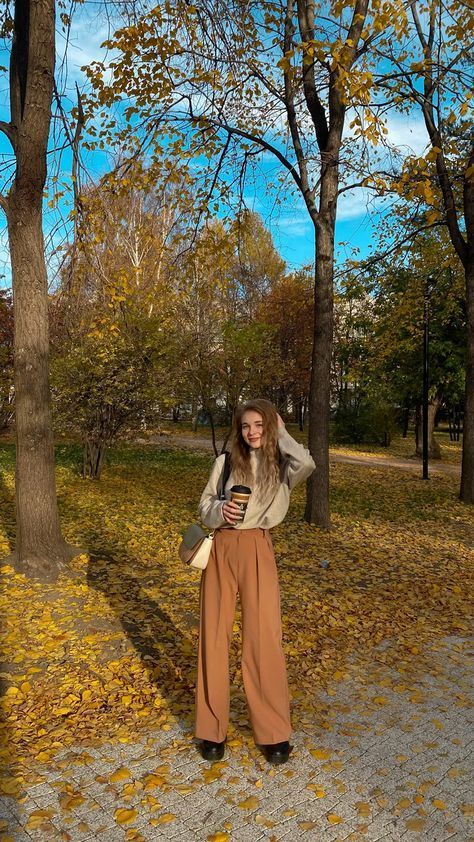 a woman standing in front of trees with leaves on the ground and wearing wide legged pants