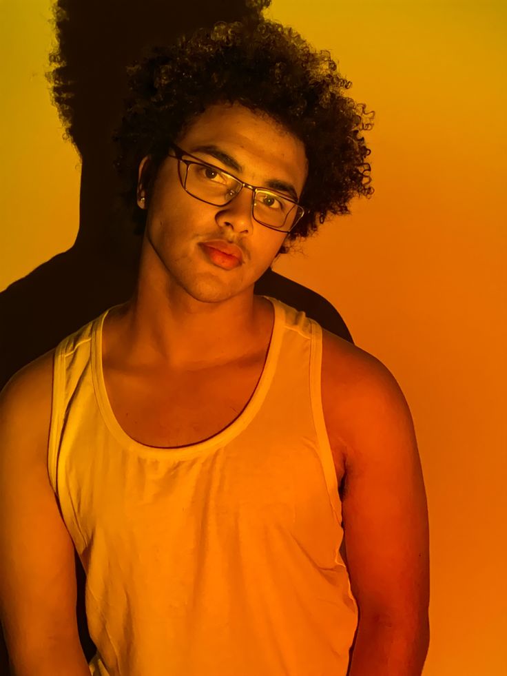 a young man with glasses standing in front of a yellow wall wearing a tank top