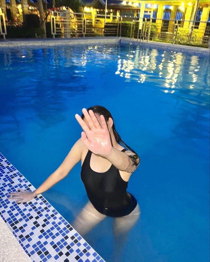 a woman in a black bathing suit standing next to a blue swimming pool at night