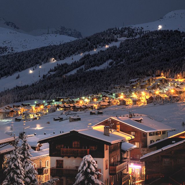 the town is lit up at night in the snow covered mountains and trees with lights on them