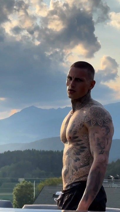 a man with tattoos standing on top of a roof in front of mountains and clouds