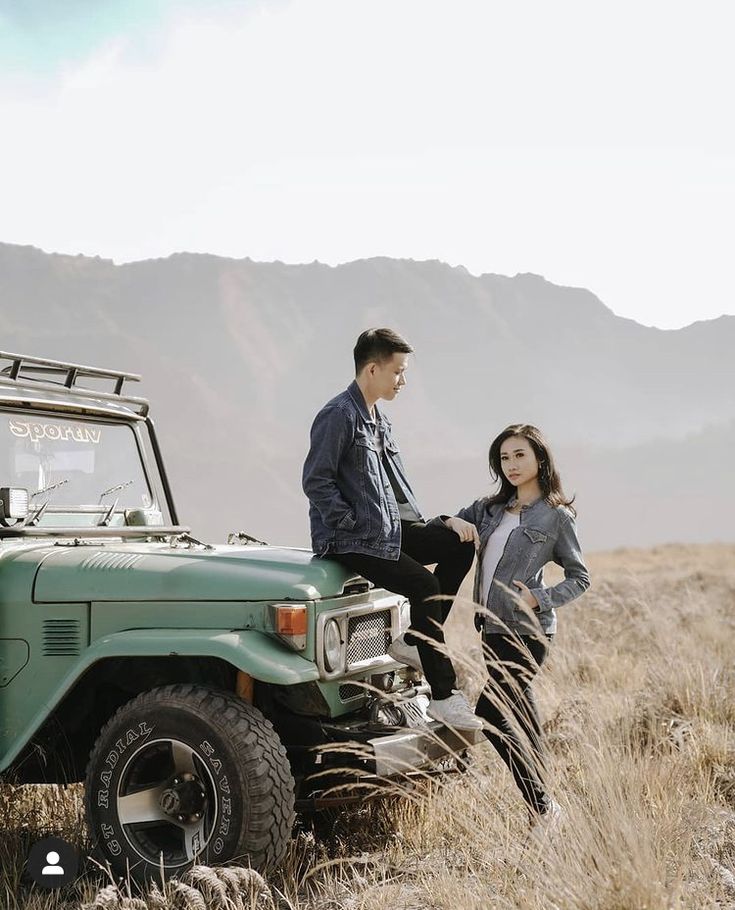 a man and woman standing next to a green jeep in the middle of a field