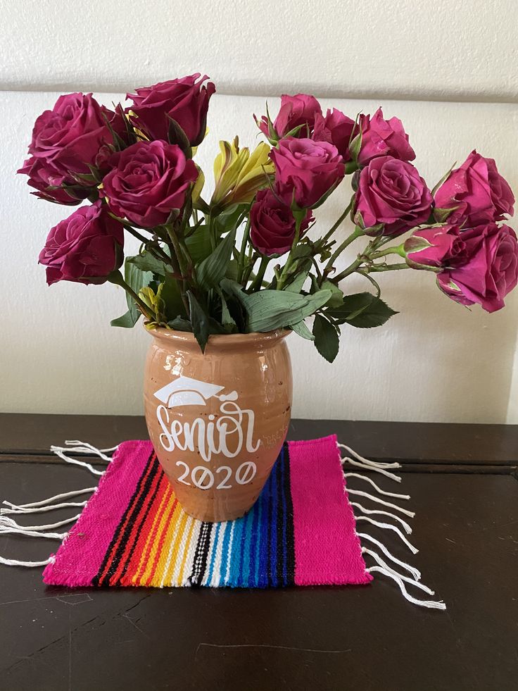 a vase filled with purple roses sitting on top of a colorful table runner next to a white wall