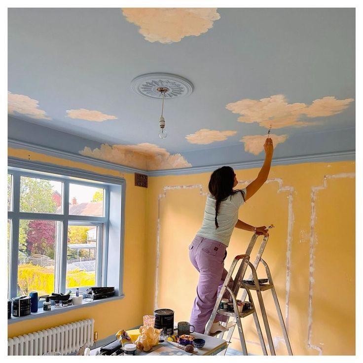 a woman standing on a ladder painting the ceiling in a room with yellow walls and clouds painted on it