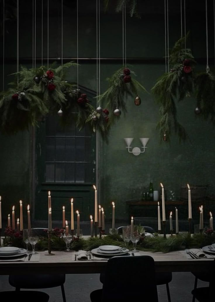 a long table with candles and greenery hanging from it's ceiling in front of a green wall