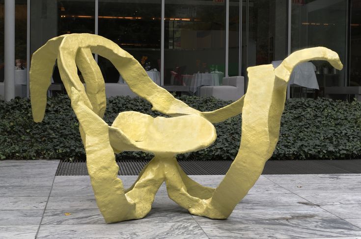 a large yellow sculpture sitting on top of a cement floor next to a building with glass windows