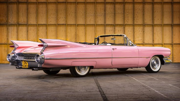 an old pink convertible car parked in a parking lot next to a wooden building with yellow walls