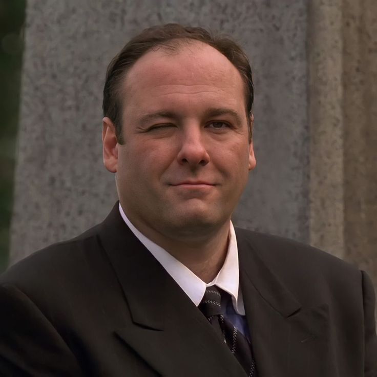 a man wearing a suit and tie standing in front of a stone wall with his arms crossed