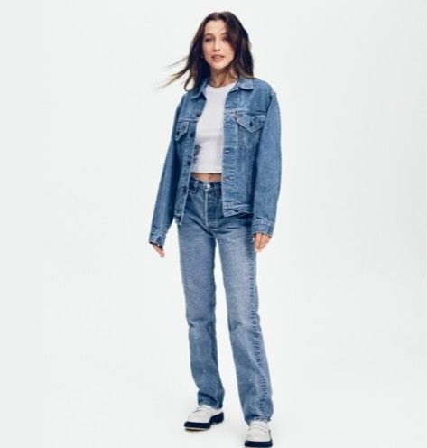 a woman standing in front of a white background wearing a jean jacket and jeans with black shoes