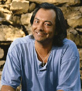 a man with long hair sitting in front of a stone wall and smiling at the camera