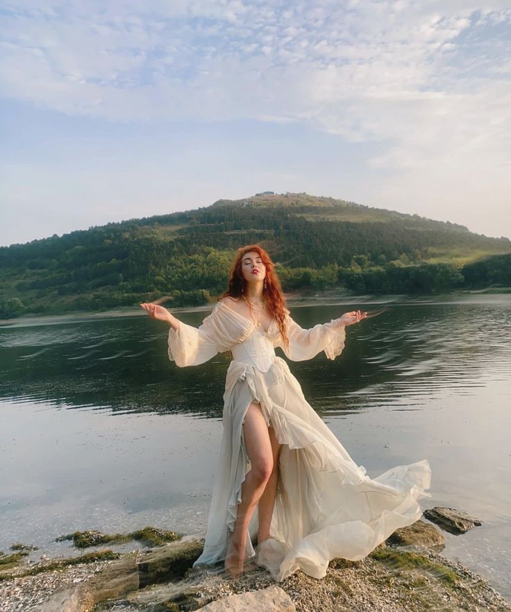 a woman in a white dress is standing on rocks by the water with her arms outstretched