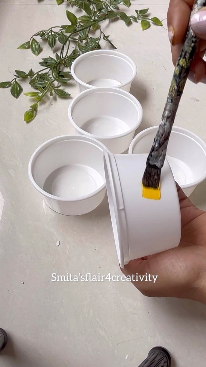 a person holding a paintbrush over four empty bowls with plants in the background on a table