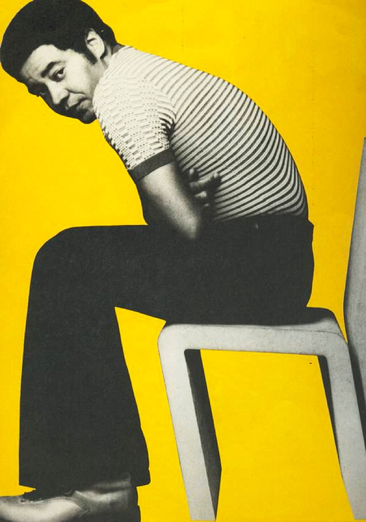 a man sitting on top of a chair in front of a yellow wall with his hand on his knees