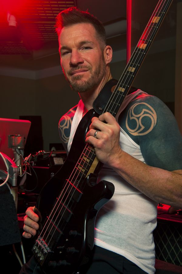 a man holding a bass guitar in his right hand and smiling at the camera with red lights behind him