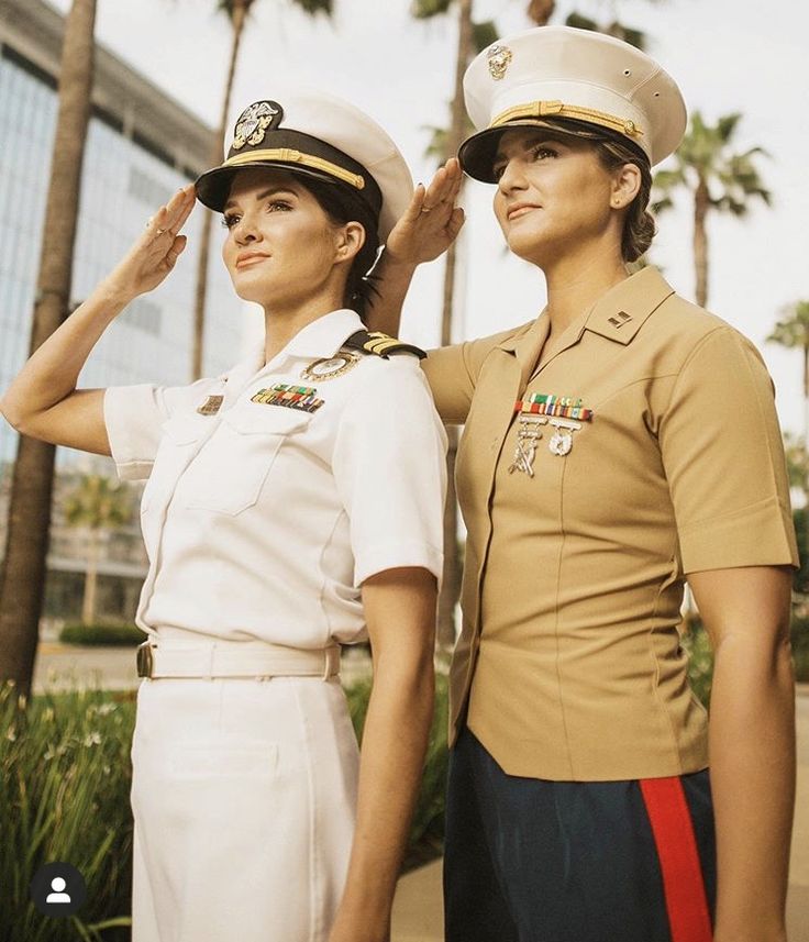 two women in uniforms standing next to each other near palm trees on the sidewalk and one woman is saluting