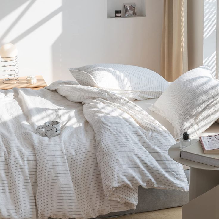 an unmade bed with white linens and pillows in a bright room, next to a window