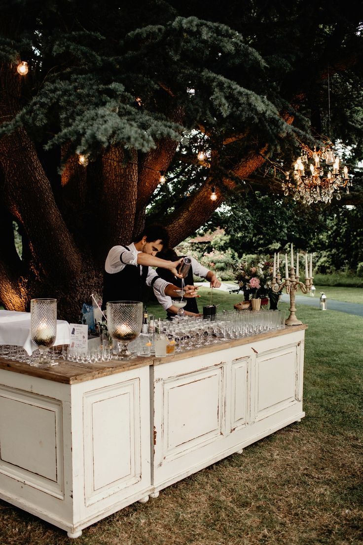 a man and woman standing at a bar with drinks on it in front of a tree