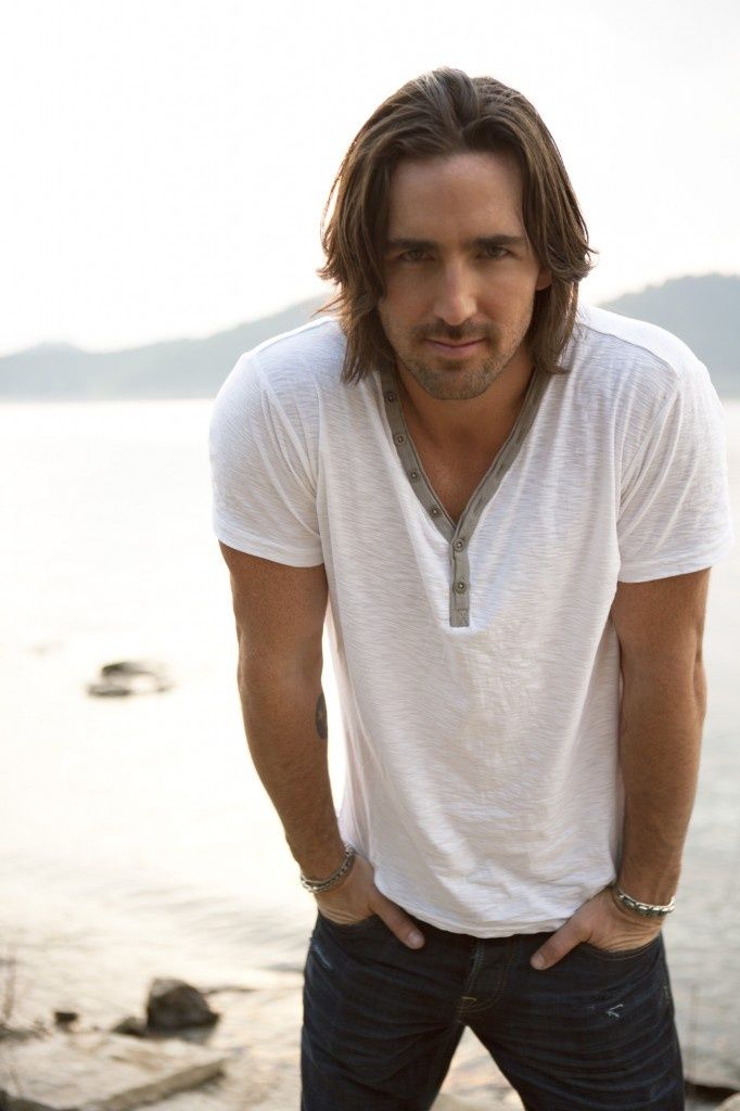 a man with long hair is standing by the water and looking at the camera while wearing a white t - shirt