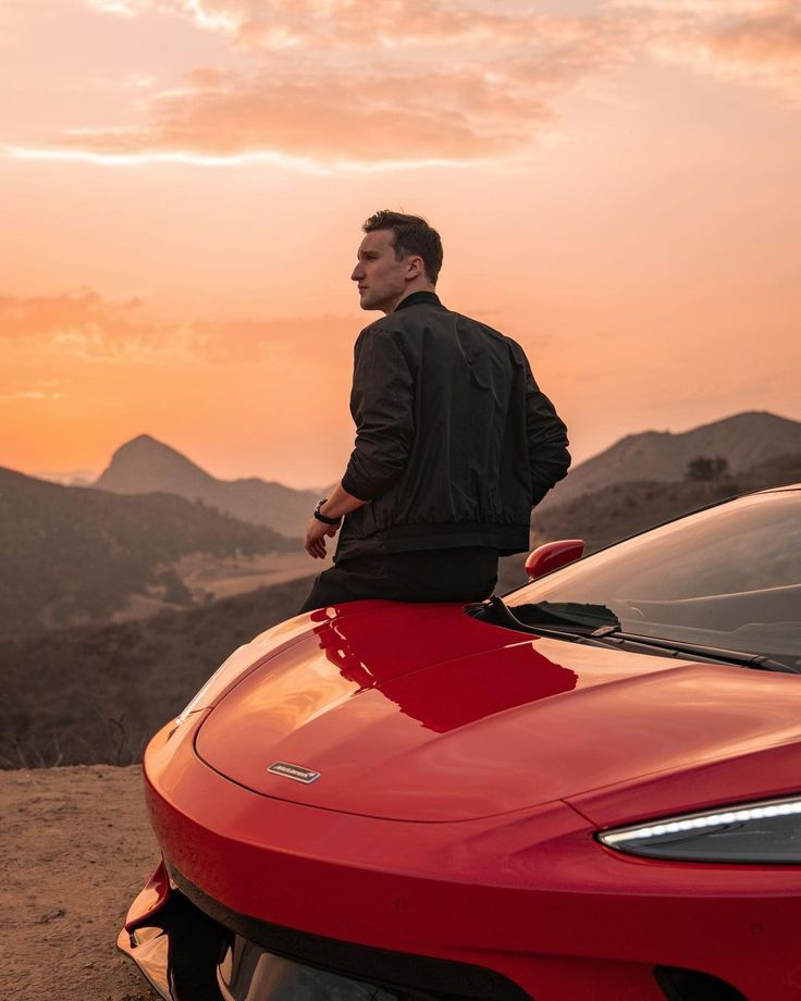 a man standing on the hood of a red sports car in front of mountains at sunset