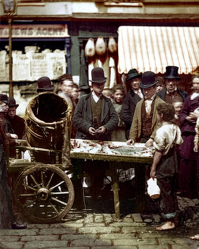 an old time photo of men and women standing around a table with food on it