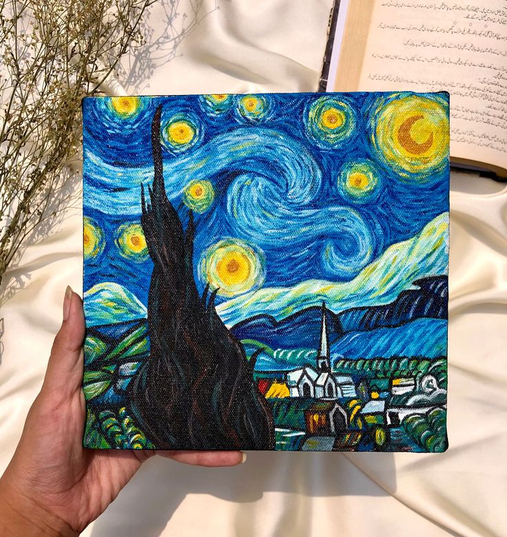 someone is holding up a starry night painting on a sheet of paper with an open book in the foreground