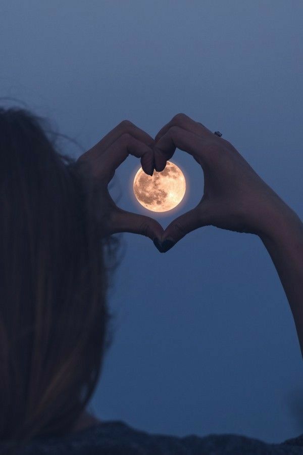 a woman making a heart shape with her hands and the moon in the sky behind her