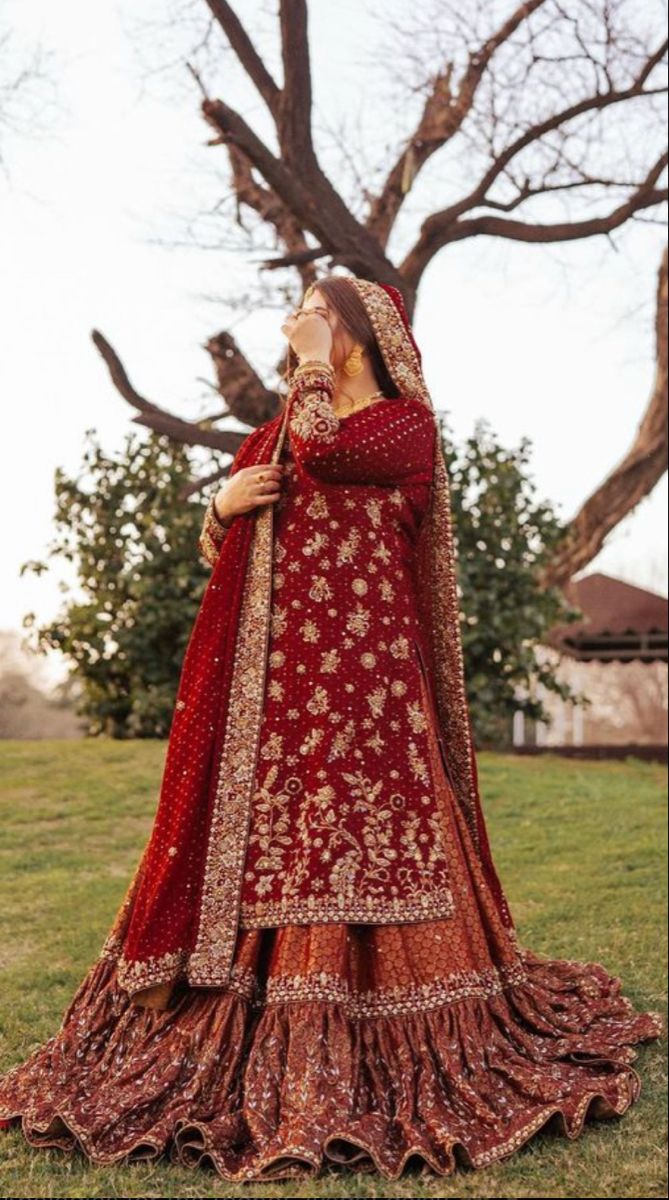 a woman in a red and gold bridal gown standing on grass with trees behind her