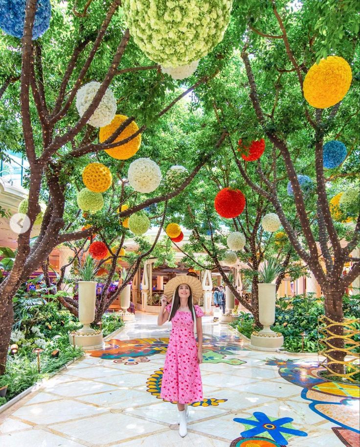 a woman in a pink dress and hat standing under trees with paper balls hanging from them