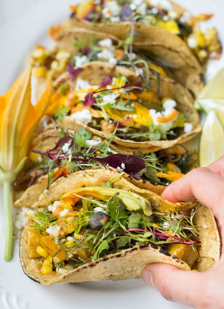 a person is holding up some tacos on a white plate with corn, lettuce and other toppings