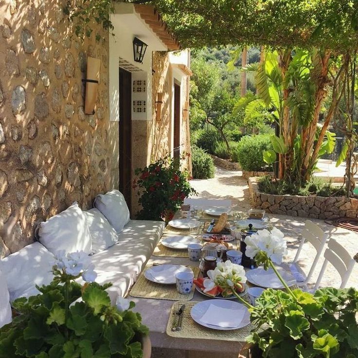 an outdoor table with plates and bowls on it in front of a stone building surrounded by greenery