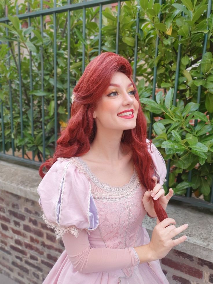 a woman with red hair wearing a pink dress and holding something in her hand while standing next to a fence