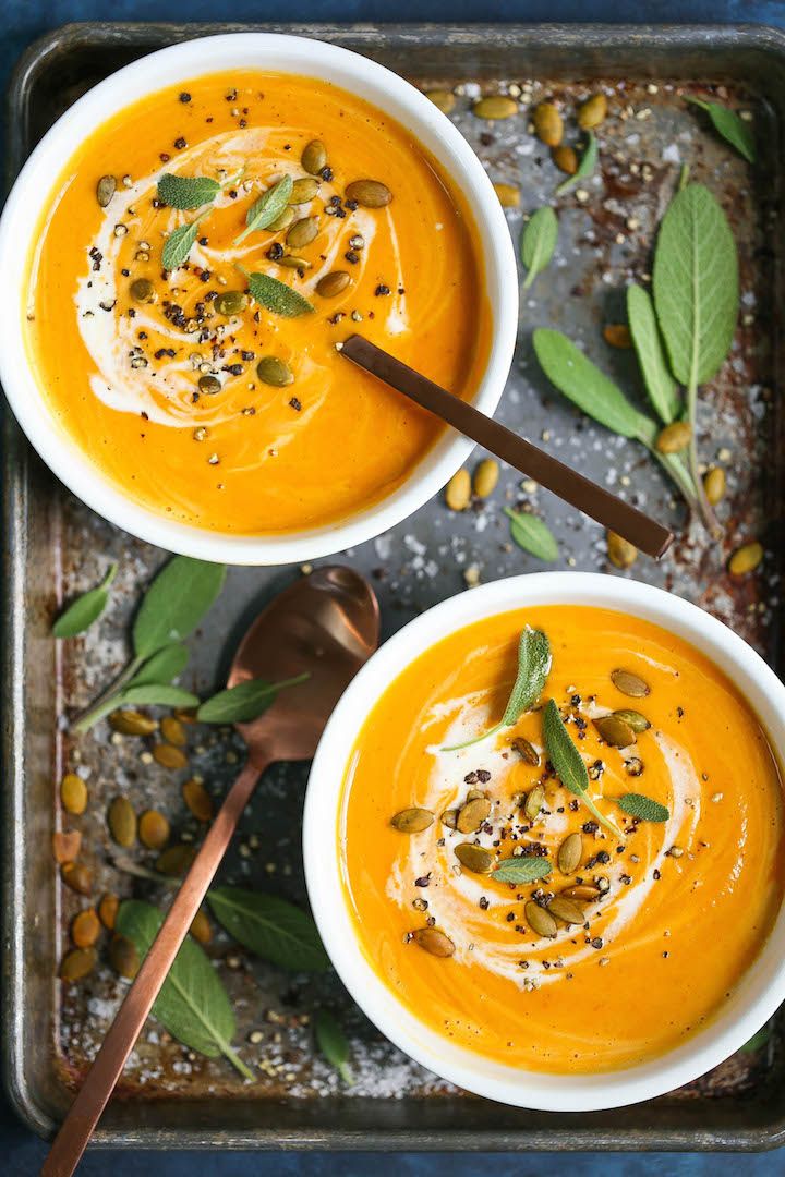 two white bowls filled with carrot soup and topped with pumpkin seeds, green leaves and spices