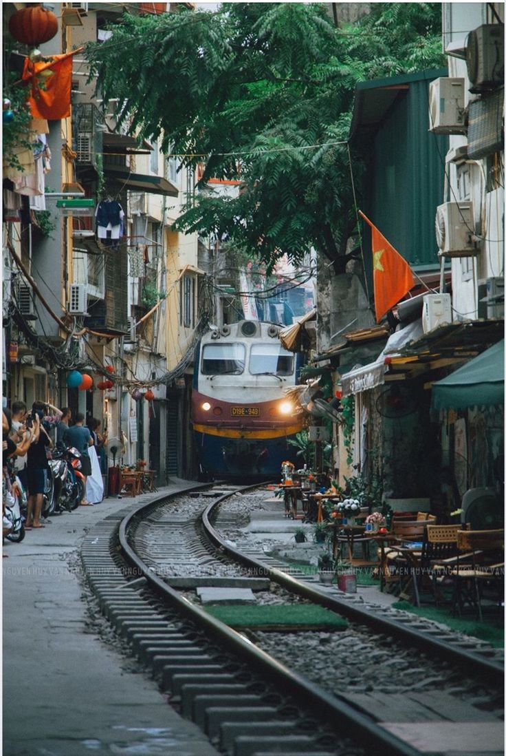 a train is coming down the tracks through an alleyway with people walking on either side