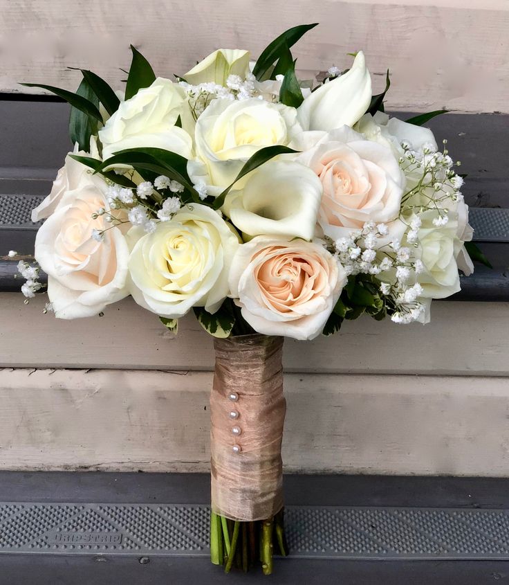 a bouquet of white and peach roses in a vase sitting on a wooden bench with greenery